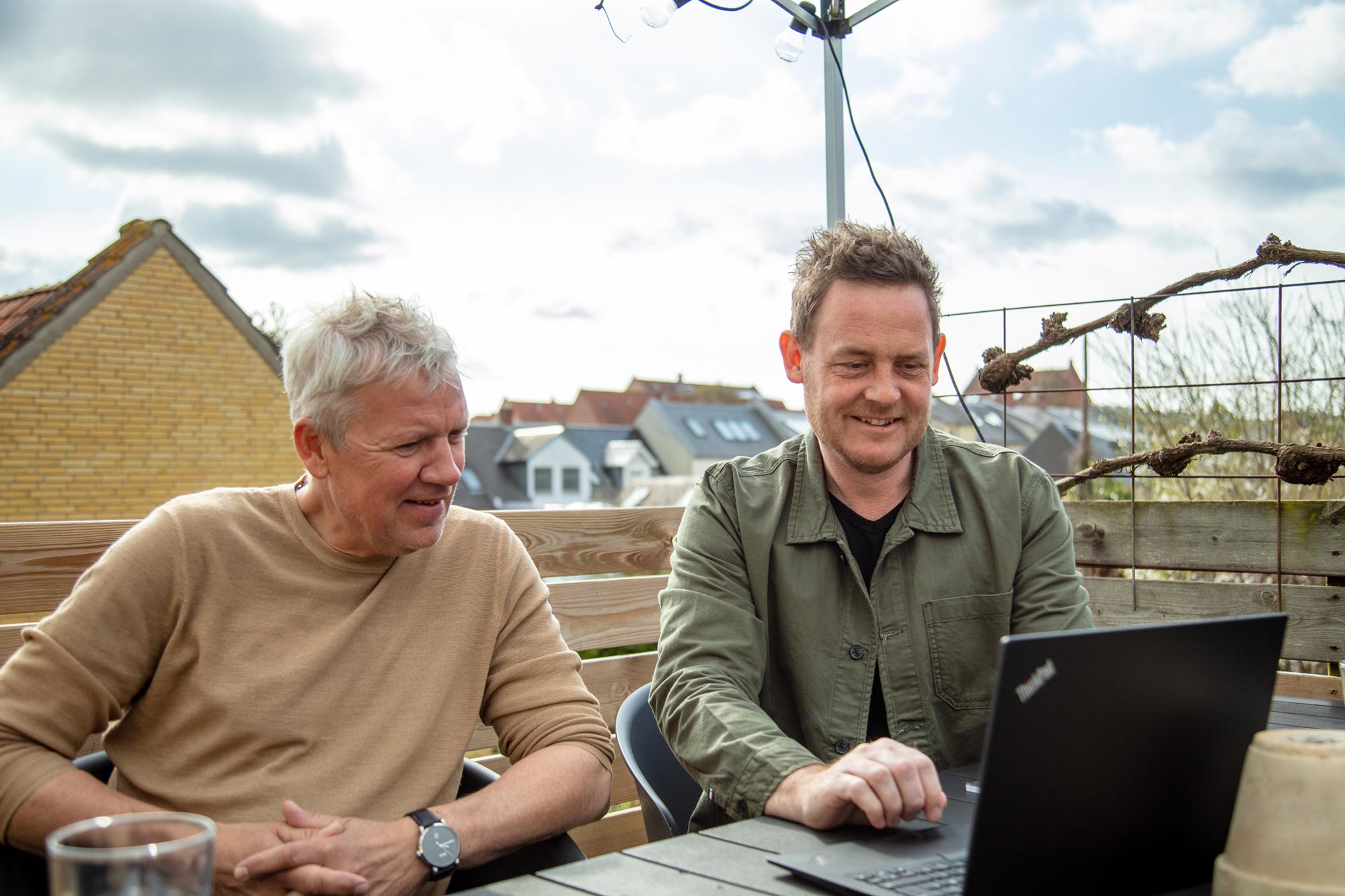 To mænd sidder ved havebord og kigger på en laptop